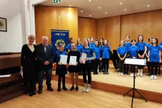 Barbara Božič in predsednik Matevž Bergant iz Lions kluba Kamnik ter tri obetavne učenke Glasbene &scaron;ole Kamnik, vsaka s posebnim priznanjem in knjižno nagrado: Anamarija Graj, Tina Bizjak in Tamara Kralj (od leve proti desni). V ozadju članice Mladinskega pevskega zbora G&Scaron; Kamnik. / Foto: Katja Dolenc